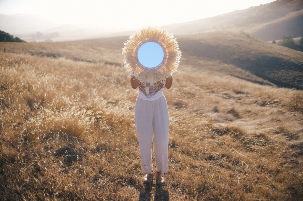 A woman holding a mirror stands in a sunlit field, reflecting the bright sky on a summer day.