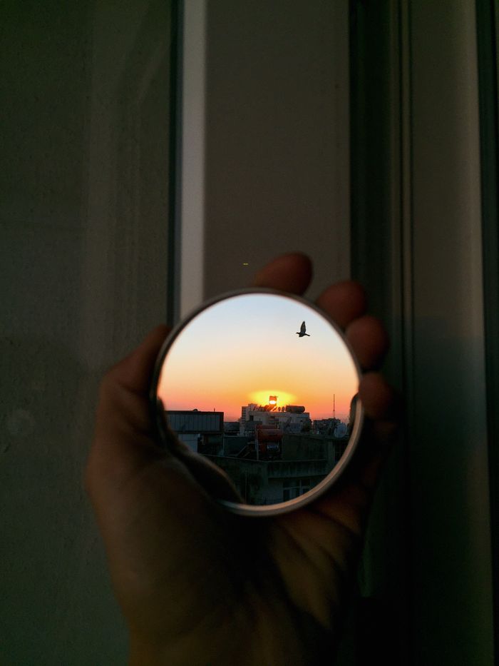 Crop anonymous person holding small round mirror with sunset and buildings reflection near window in evening